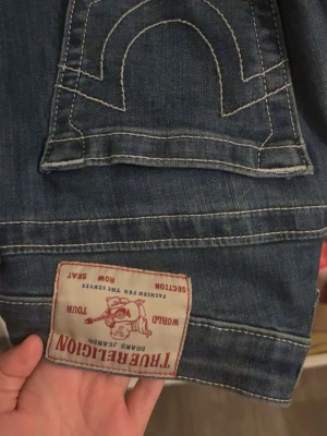 Blå True Religion jeans med kontrastsöm - Säljer ett par klassiska blå jeans från True Religion med deras ikoniska vita kontrastsömmar och stor bakficka. Jeansen har en tydlig logopatch bak och är tillverkade i denim med snygg tvätt. Dem är nersydda så dem passar till kanske 1.59 cm, dem har hål mellan benen därför är det ett billigt pris. Lätt att sy mellan benen. 