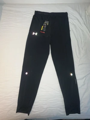 Under Armour byxor - Säljer ett par svarta träningsbyxor från Under Armour med reflekterande logga på vänster lår och reflexdetaljer på benen. Byxorna har elastisk midja och är tillverkade i ett lätt, snabbtorkande material som passar perfekt för löpning eller gym. Byxorna är storlek L men passar även M. Aldrig använda! Pris kan diskuteras!