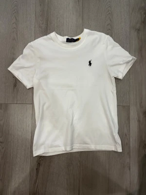 Vit t-shirt från Polo Ralph Lauren - Klassisk vit t-shirt från Polo Ralph Lauren med korta ärmar och rund halsringning. T-shirten har den ikoniska svarta polospelaren broderad på bröstet. Perfekt basplagg i mjuk bomull som funkar till allt.