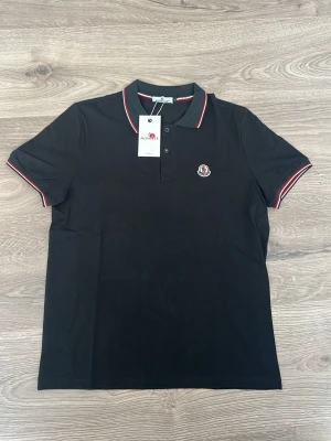 Svart pikétröja från Moncler - OBS. Loggan är väl dåligt gjort eller helt okej. Därav låga pris. Kan passa även M. Svart pikétröja från Moncler med klassisk krage och knäppning framtill. Tröjan har röda och vita detaljer på krage och ärmslut samt Moncler-logga broderad på bröstet. Tillverkad i bomull för en bekväm känsla.