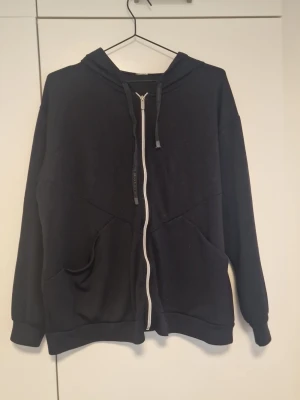 Svart hoodie med dragkedja - En enkel svart hoodie med dragkedja framtill och två fickor. Den har huva med dragsko och ribbade muddar vid ärmar och nederkant. Perfekt för en avslappnad stil och funkar året runt. Materialet känns mjukt och bekvämt, troligen bomullsblandning.