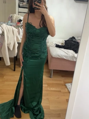 Grön långklänning med slits - Elegant grön långklänning med smala axelband och broderade detaljer upptill. Klänningen har en figurnära passform, slits framtill och är tillverkad i ett glansigt material som ger en lyxig känsla. Perfekt för speciella tillfällen.