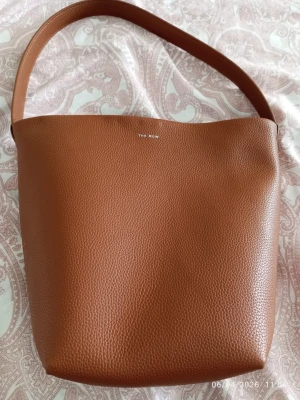The Row Park Tote Medium Brown - Snygg brun bucket bag från The Row i mjukt, kornigt läder. Väskan har en minimalistisk design med bred axelrem och diskret logga framtill. Perfekt för dig som gillar stilrena accessoarer med lyxig känsla.