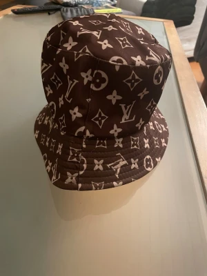 bucket hat, vändbar - Snygg vändbar bucket hat  . Ena sidan är brun med klassiskt monogrammönster i vitt, andra sidan är helsvart med stor -logga framtill. Hatten är i tyg och har en avslappnad passform. Perfekt accessoar för att lyfta din stil.