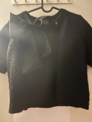 Svart kofta med stor rosett - Snygg svart kofta med korta ärmar och en stor dekorativ rosett vid halsen. Koftan har rund halsringning och knäppning framtill. Perfekt för dig som vill ha en stilren men ändå unik look.