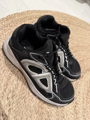 Svarta sneakers med silvriga detaljer  - Svarta sneakers med silvriga och vita detaljer, sportig design och meshpartier för extra ventilation. Skorna har en tjock vit sula och svarta skosnören. Perfekta för en streetwear-look eller till vardagsoutfiten.