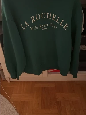Grön sweatshirt - Mörkgrön sweatshirt från La Rochelle med trycket 'Vélo Sport Club' i vitt framtill. Tröjan har rund halsringning, långa ärmar och en avslappnad passform. Perfekt för en chill och sportig stil.