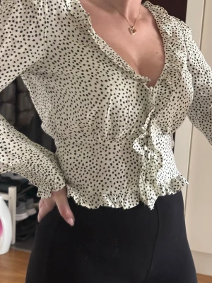 Prickig blus med volang från BikBok - Supersöt vit blus med svarta prickar från BikBok. Blusen har långa ärmar, volangkant längs knäppningen och nederkanten samt en djup v-ringning. 