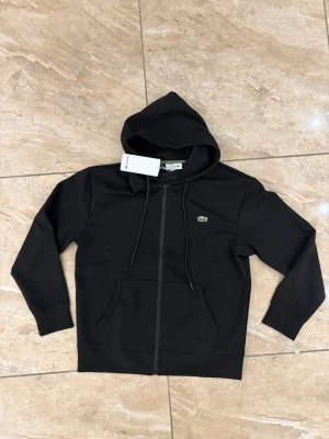 Svart zip-hoodie från Lacoste - Svart hoodie med dragkedja från Lacoste. Klassisk design med huva och dragsnören, två fickor framtill och den ikoniska gröna krokodilloggan på bröstet. Tillverkad i mjukt bomullsmaterial för en skön och avslappnad stil.