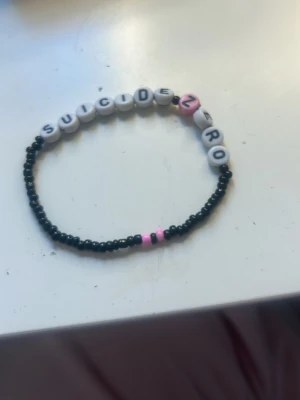Pärlarmband med text SUICIDE ZERO - Ett handgjort armband med svarta små pärlor och vita bokstavspärlor som bildar texten SUICIDE, skänker pengarna till organisationer som tar hand om barn och unga. Perfekt accessoar för dig som vill bära ett viktigt budskap.