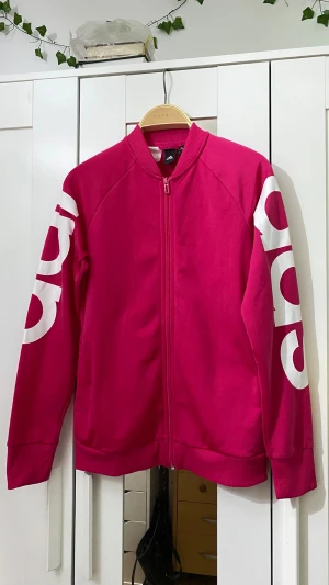 Rosa Adidas bomberjacka med tryck - Snygg rosa bomberjacka från Adidas med vit logotyp-tryck längs båda ärmarna. Jackan har dragkedja framtill, ribbade muddar och klassisk krage. Perfekt för dig som vill sticka ut med sportig stil.