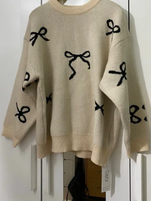 Beige stickad tröja med rosett-mönster - Snygg beige stickad tröja från DAZY med svarta rosetter som mönster. Tröjan har en avslappnad passform, rund halsringning och ribbade muddar vid ärmslut och nederkant. Perfekt för dig som gillar trendiga detaljer och oversized stil.