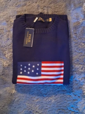 Mörkblå stickad tröja Polo Ralph Lauren - 3 Mörkblå stickad tröja från Polo Ralph Lauren med amerikansk flagga framtill. Tröjan har rund halsringning och ribbstickade muddar. Perfekt för dig som gillar klassisk och stilren design.