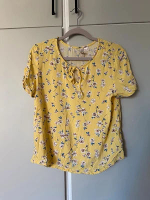 Gul blommig blus  - Jättefin vårig topp/tshirt. Finns detaljer såsom snöre fram samt knäppe på ärmens sida. Använd vid ett tillfälle förutom det oanvänd!