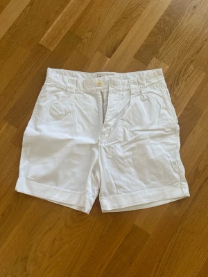 Vita shorts från Gant med hög midja - Klassiska vita shorts från Gant med hög midja och bälteshällor. Tillverkade i mjuk bomull med knapp och dragkedja framtill. Perfekta för varma dagar och enkel att matcha med olika stilar.