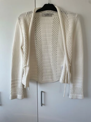 Vit stickad kofta från Clara - Snygg vit stickad kofta från Clara med långa ärmar och knytband i midjan. Koftan har ett öppet hålmönster längs ärmar och fram- och baksida, vilket ger en luftig och trendig look. Perfekt att slänga över en topp för en chill stil.