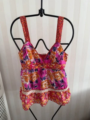 Blommig linnetopp med spetsdetalj - Färgglad linnetopp med blommigt mönster i rosa, orange och lila toner. Toppen har breda axelband, v-ringning och en volangkant nertill. Dekorerad med spetsdetalj och smockad rygg för extra komfort. Perfekt för varma dagar.