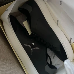 Svarta Axel Arigato sneakers med fågel. - Säljer ett par svarta sneakers från Axel Arigato med vit sula och en snygg broderad fågel på sidan. Skorna har en clean och stilren design med lågt skaft och svarta skosnören. Dock lite slitna inuti men inget som syns på utsidan, slitheten märks inte av när du använder dem.