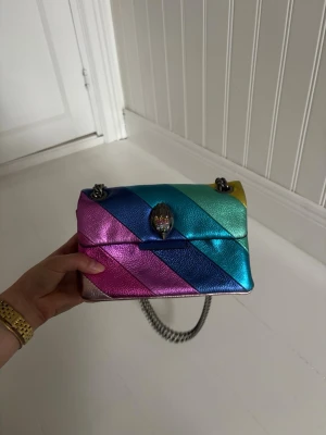 Regnbågsfärgad handväska Kurt Geiger - Snygg handväska från Kurt Geiger i regnbågsfärgat metallicläder med diagonalrandigt mönster. Väskan har en silverfärgad kedjerem och ikoniskt örnhuvud som spänne. Perfekt nu till sommaren! Nypris 2445🌸🥰