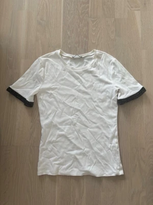Vit t-shirt med svarta spetsdetaljer - Säljer en vit t-shirt med svarta spetsdetaljer vid ärmsluten. T-shirten har rund halsringning och korta ärmar. Materialet känns mjukt och passar perfekt till en enkel men snygg look. Storlek M men passar absolut en S, brukar ha det vanligtvis! 