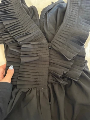 Svart blus - Snygg svart blus med breda plisserade volanger framtill som ger en cool och unik look. Blusen har en v-ringad hals och är tillverkad i ett mjukt tyg som faller fint. Perfekt för dig som vill sticka ut lite extra. Oanvänd. 