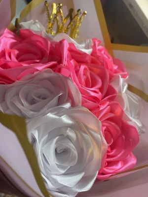 Bukett med konstgjorda rosor och krona - Säljer en bukett med konstgjorda rosor i rosa och vitt, gjorda av glansigt tyg. Buketten är dekorerad med en liten glittrig guldfärgad krona och inslagen i papper med guldkant. Perfekt som present eller dekoration.