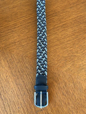 Blått flätat bälte från ONEWear - Snyggt flätat bälte från ONEWear i olika blå och grå toner. Bältet har ett randigt mönster och detaljer i mörkblått skinn vid spännet. Perfekt accessoar för att lyfta din outfit. 