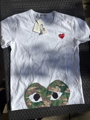 Vit Comme des Garçons PLAY t-shirt - Vit t-shirt från Comme des Garçons PLAY med det klassiska röda hjärtat på bröstet och ett stort camo-hjärta med ögon längst ner. Kortärmad och i mjuk bomull, perfekt för en chill och trendig look.