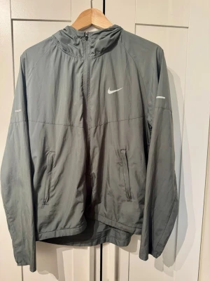 Grå Nike miler vindjacka  - Snygg grå Nike Miller vindjacka med luva och detaljer på armen samt skiner dem och Nike logan i mörkret. I storlek S 