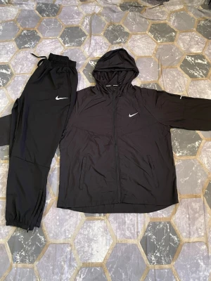 Nike miler  - Säljer ett svart träningsset från Nike med matchande byxor och jacka. Jackan har dragkedja, huva och vita Nike-loggor på bröstet och byxan. Materialet är lätt och passar perfekt för träning eller chill. Byxorna har resår i midjan och vid benslut.
