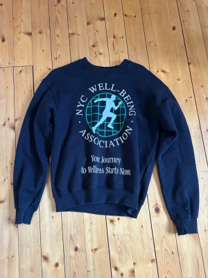 Svart sweatshirt med tryck - Cool svart sweatshirt med stort tryck framtill: 'NYC WELL-BEING ASSOCIATION' och en grafisk figur i turkos. Rund hals, ribbade muddar och avslappnad passform. Perfekt för en streetwear-look och enkel att matcha med jeans eller joggers. Från Carlings i storlek S.