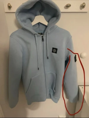 Stone island tröja  - Snygg ljusblå hoodie med dragkedja och huva. Tröjan har två fickor framtill, justerbara snören i huvan och en svart patch på bröstet. Tillverkad i mjukt material som känns skönt mot huden. Perfekt för en avslappnad och trendig look.