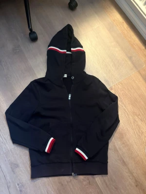 Svart hoodie från Moncler med ränder - Snygg svart hoodie från Moncler med dragkedja framtill och huva. Detaljer i form av röda och vita ränder på huvan och vid ärmsluten ger en sportig vibe. Praktiska fickor på magen och mjukt material som känns skönt mot huden. ÄKTA Bluetooth funkar mot yttre loggan