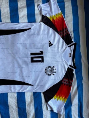Tysklands matchtröja - Musiala  - Vit fotbollströja från Tysklands landslag med nummer 10 på bröstet. Tröjan har svarta detaljer och mönster i rött, gult och svart på axlarna. Tysklands landslagsemblem med fyra stjärnor finns på bröstet. Perfekt för fotbollsfans! Storlek L men passar även som M. Endast testad en gång.