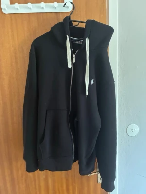 Svart hoodie från Polo Ralph Lauren - Säljer en svart hoodie från Polo Ralph Lauren med dragkedja, vit logga på bröstet och vita snören i huvan. Hoodien har två fickor framtill och är gjord i mjukt material som känns skönt mot huden. Perfekt för en avslappnad och stilren look.