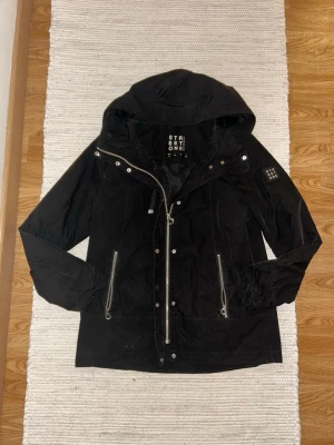Svart vindjacka från Street One - Snygg svart vindjacka från Street One med huva, dragkedja och tryckknappar framtill. Jackan har två framfickor med dragkedja och ett meshfoder på insidan. Patch med logga på ärmen och justerbara muddar. Perfekt för blåsiga dagar. Storlek 34 / S. Användes förra sommaren, ge prisförslag 