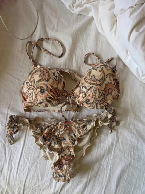 Beige paisleymönstrad bikini - Säljer en beige bikini med paisleymönster i blått, rosa och orange. Överdelen har vadderade kupor, knytning fram och smala axelband. Underdelen har volangkant och knytband i sidorna. Materialet känns mjukt och stretchigt, perfekt för stranden eller poolen.