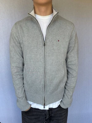 Ljusgrå stickad ziptröja Tommy Hilfiger XS - Detta är en kofta från Tommy Hilfiger i storlek XS med normal passform. Modell på bild är ca 180, 66kg. Plagget är i bra skick utan några defekter. Hör gärna av dig vid intresse eller frågor! 