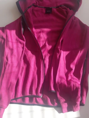 Mörkrosa hoodie med dragkedja från Peak performance  - Snygg mörkrosa hoodie med dragkedja framtill och huva. Tillverkad i ett mjukt och stretchigt material som känns skönt mot huden. Perfekt för chill dagar eller när du vill ha en färgklick i din outfit. Svarta detaljer längs dragkedjan och huvan.