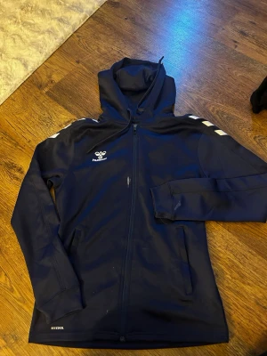 Marinblå Hummel hoodie med dragkedja - Snygg marinblå hoodie från Hummel med dragkedja framtill och huva. Klassiska vita Hummel-pilar på axlarna och logga på bröstet. Tillverkad i ett funktionsmaterial som andas, perfekt för träning eller uppvärmning. Tröjan är som ny i skicket men har fläckar (se bilder) därav priset