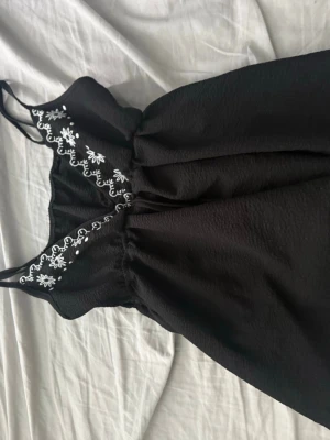 Svart nattlinne med spetsdetaljer - Säljer ett svart nattlinne med tunna axelband och v-ringning. Dekorerat med vita broderade blommor längs ringningen och lätt rynkad midja för en bekväm passform. Stolek S aldrig använd