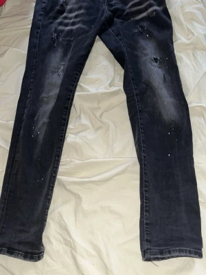 Svarta distressed jeans från Dsquared2 - Säljer ett par svarta jeans från Dsquared2 med slitningar, hål och färgstänk för en edgy look. Jeansen har smal passform och klassisk femficksdesign. Perfekta för dig som gillar streetstyle och vill sticka ut. Passar folk som är kanske 183cm och 65kg eller mer lite stora för mih