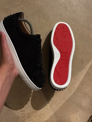 Louboutin skor  - Säljer ett par svarta sneakers med coola svarta nitar på tå och häl. Skorna har en vit sula och en röd undersida som sticker ut. Materialet ser ut att vara mocka eller liknande tyg. Klassisk låg modell med svarta skosnören och snygg kontrast mellan färgerna. Ingen box tillkommer.