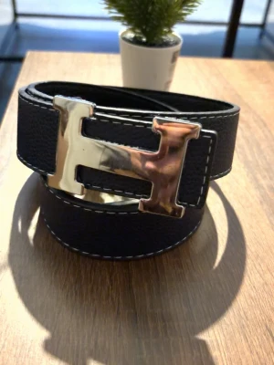 Bälte  - Snyggt svart bälte med ett stort, blankt H-format metallspänne. Bältet har vita kontrastsömmar längs kanterna och är gjort i ett strukturerat material som liknar skinn. Perfekt för att lyfta vilken outfit som helst.