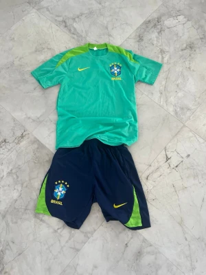 Brasilien fotbollsträningsset Nike - Säljer ett Brasilien fotbollsträningsset från Nike. Tröjan är ljusgrön med limegröna detaljer och har Brasil-logga och Nike-swoosh på bröstet. Shortsen är marinblå med limegröna sidor och har Brasil-märke och Nike-logga. Perfekt för träning eller match!
