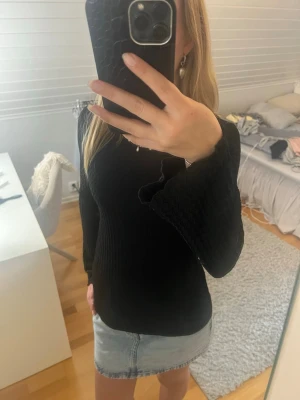 Svart tröja med utsvängda ärmar - Snygg svart tröja med vågig struktur och utsvängda ärmar. Tröjan har rund halsringning och är tillverkad i ett stretchigt material som sitter skönt på kroppen. Perfekt för dig som vill ha en stilren men ändå unik look.