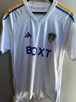 Leeds United matchtröja vit - Säljer en vit Leeds United matchtröja med blå och gula detaljer på axlarna. Tröjan har klubbmärke och Adidas-logga på bröstet samt BOXT som sponsortryck. På ryggen står det 'Summerville' och nummer 10 i blått. Tillverkad i lätt och ventilerande material.