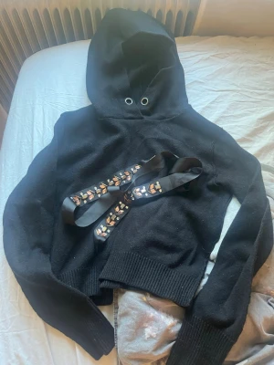 Svart cropped stickad  hoodie med snörning - Säljer en svart cropped hoodie med huva och cool snörning framtill. Hoodien har långa ärmar och en avslappnad passform som passar perfekt till jeans eller tights. Materialet känns mjukt och bekvämt mot huden. Passar mig som är xs-s, har tagit bort snörerna men skickar med dom för de går att sätta på igen