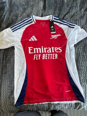 Arsenal matchtröja röd/vit Adidas - Säljer en officiell Arsenal matchtröja från Adidas i rött, vitt och marinblått. Tröjan har klubbmärke och sponsortryck framtill, samt klassiska Adidas-ränder på axlarna. Tillverkad i lätt och ventilerande material, perfekt för fotbollsträning eller match.