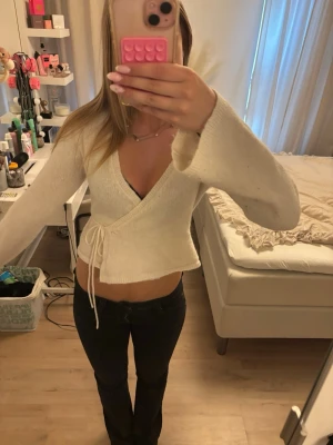 Vit omlottstickad tröja - Säljer en vit stickad omlottopp med långa ärmar och knytband i midjan. Tröjan har en djup v-ringning och är gjord i mjukt stickat material. Perfekt att styla med jeans eller kjol för en trendig look.